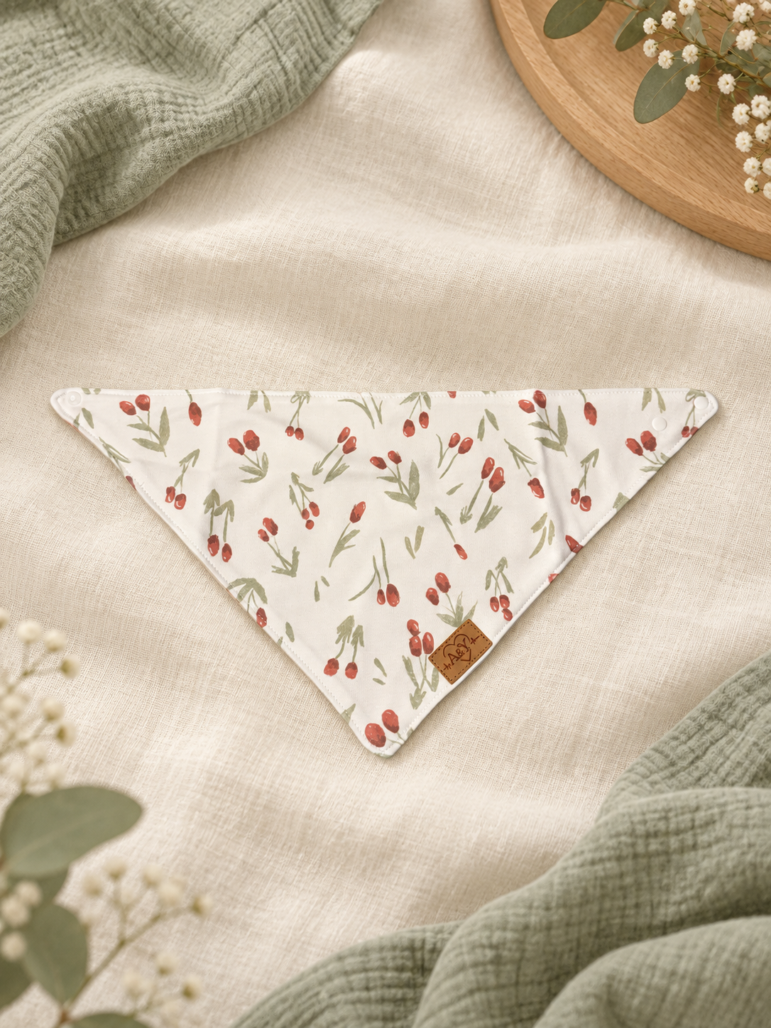 MINUS natural cotton bandana bib in the Red Flowers pattern, laid flat on a cream and sage background.