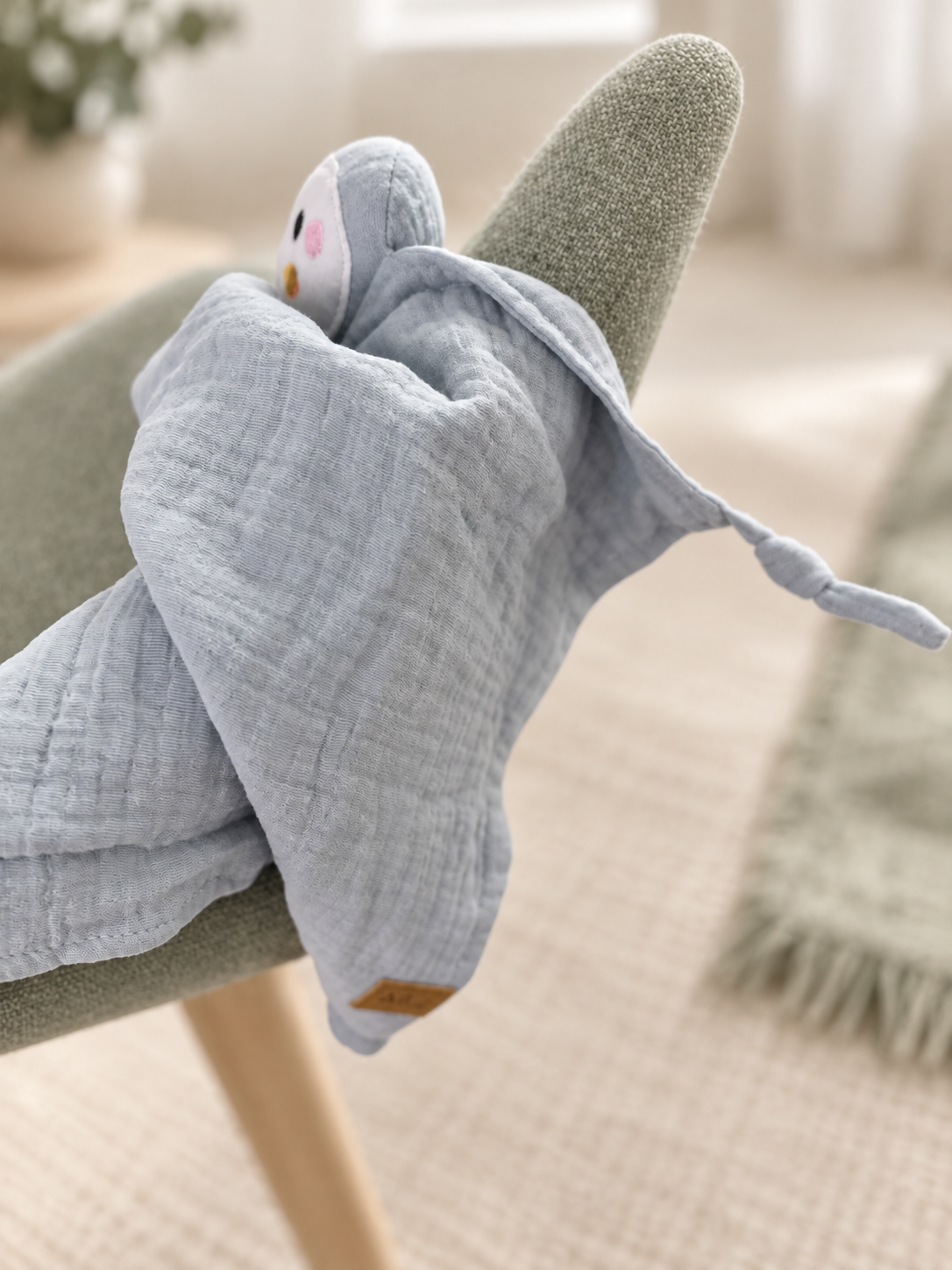 Doudou MINUS pingouin bleu-gris drapé sur une chaise dans un décor sauge.