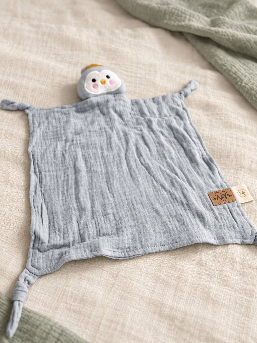 Doudou MINUS pingouin présenté de biais, avec son visage brodé et ses coins noués.