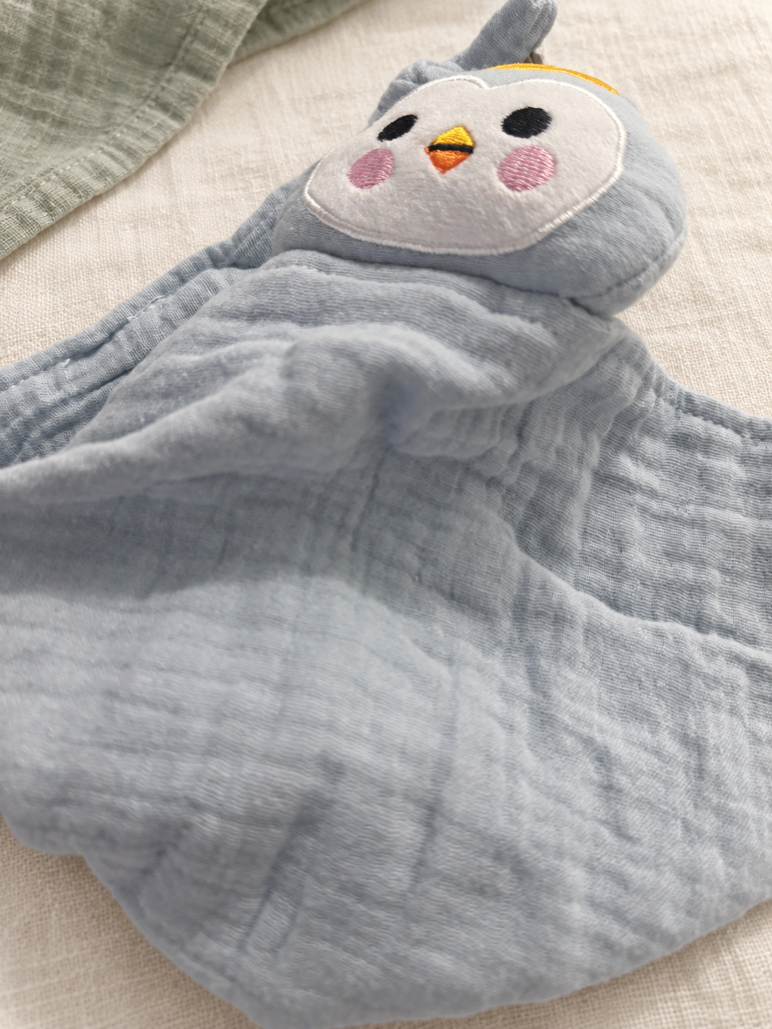 Gros plan du visage brodé du doudou MINUS pingouin et de son tissu bleu-gris.
