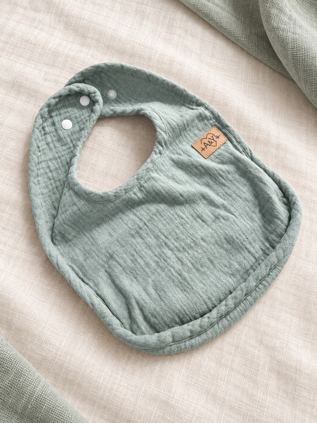 Bavoir MINUS sauge présenté de biais pour montrer sa forme et son tissu.